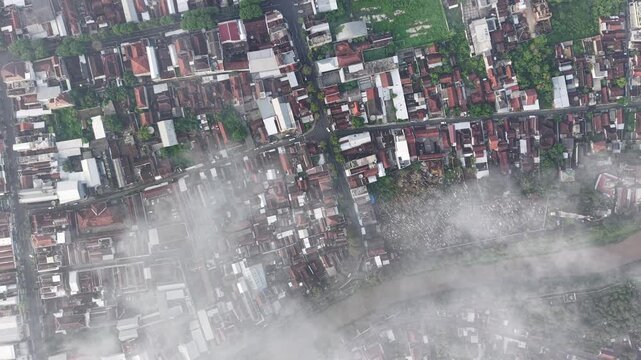 An aerial scene of a crowded city block with red rooftops, narrow streets, and a river along the edge, partially veiled by morning fog. Evokes urban density, city life, and misty atmosphere.