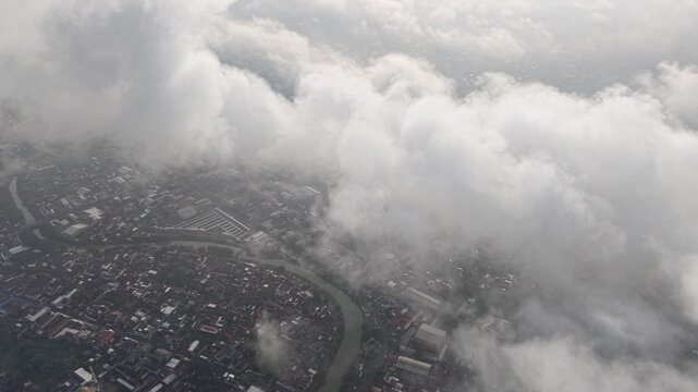 An aerial view of a dense urban area with a winding river, bridges, and a network of streets, partially veiled by fluffy clouds. The scene captures city life from above on a bright day.