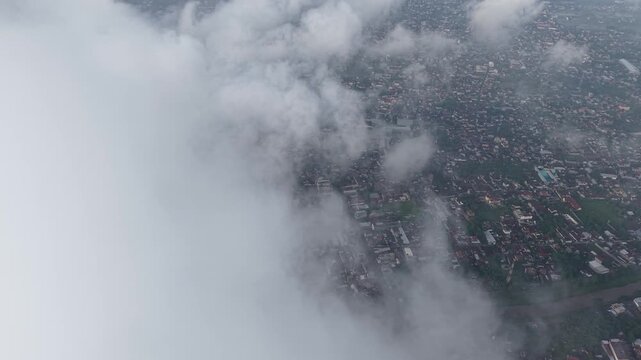 An aerial photograph of a sprawling city partially veiled by thick clouds and fog. Buildings and rooftops hint at urban life beneath a moody, atmospheric sky, conveying distance and mystery from above
