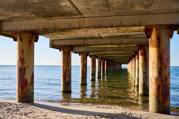 Rusted support beams under Baltic Sea pier with calm water and sandy beach
