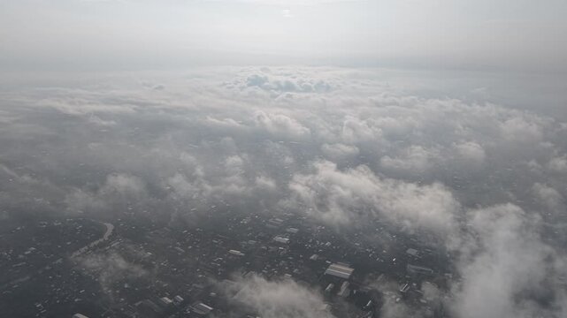 An aerial shot shows a city beneath a soft cloud layer, framed by a pale sky. The image conveys travel, distance, and the quiet majesty of flight.