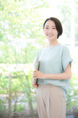 綺麗なオフィスの緑背景でオフィスでパソコンを持つミドルの笑顔のカメラ目線の女性　春夏のオフィスカジュアル　縦