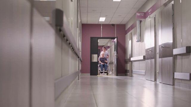 Stab shot of middle-aged Caucasian female nurse in scrubs assisting senior woman in wheelchair, walking along hospital corridor in modern clinic
