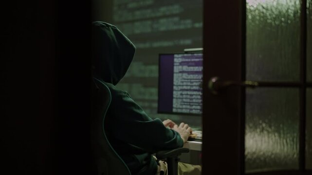 Hooded person typing on computer keyboard in dark room, focusing on screen displaying lines of code with additional green digital text projected onto wall. Concept of cyber attack, malware.