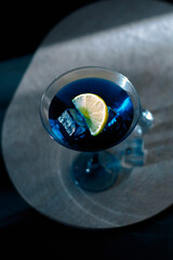 Electric blue cocktail in elegant martini glass with ice cubes and fresh lime slice. Moody dramatic lighting, sophisticated top-down view &mdash; perfect for luxury bar, party and summer refreshment concept