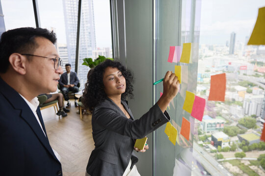 Female employee writing on adhesive note standing next to manager at office
