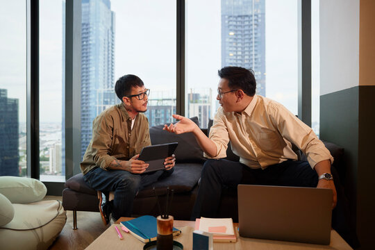 Male manager discussing strategies with young colleague while sitting on sofa at office