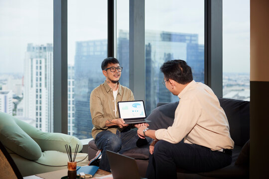 Young male entrepreneur showing chart diagram to manager on digital tablet while sitting on sofa at office