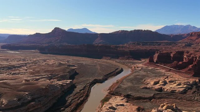 A high aerial perspective shows an opaque amber river snaking through deep red rock basins and ancient sandstone canyons. Snowy mountain peaks sit on the far horizon of this scifi Utah desert.