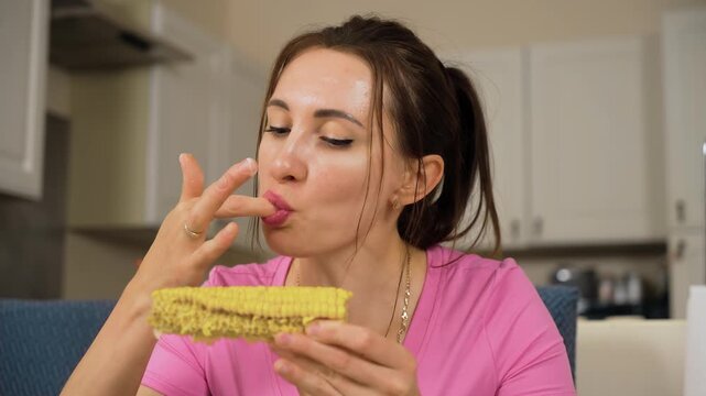Female licking finger after eating corn cob at table. Woman enjoying taste of maize while holding cob in hand. Girl savoring sweetcorn and cleaning hand during meal in kitchen