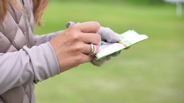 A woman checks her golf round results on the scorecard.. She is wearing a gray jacket and gloves