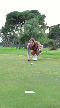 A woman is kneeling down on a golf course, preparing to hit a golf ball with a putter
