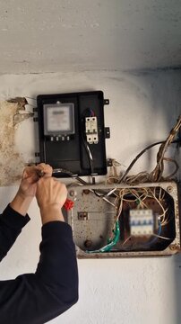Vertical, close-up of an electrician's hands stripping insulation from an electrical wire using a stripper tool. An old, exposed circuit breaker box and energy meter are in the background.