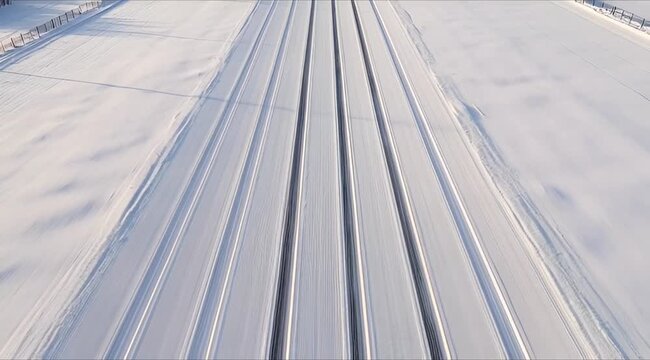 A high-angle aerial view of multiple parallel tracks etched into a vast, pristine white snowfield, stretching towards the horizon under a clear sky.