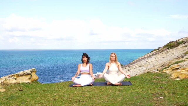 Diverse female friends practicing yoga meditation together on a scenic cliffside with a beautiful ocean view, focusing on mindfulness, wellness, and finding inner peace in nature