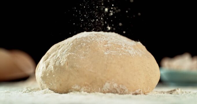 Static shot of a leavened dough loaf isolated on black background with falling flour in super slow motion, 1000 fps. Concept of baking, artisan bread making and dough preparation.