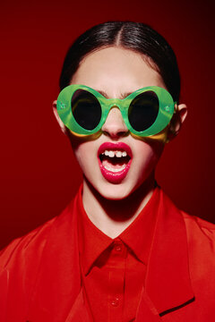 Young woman with oversized green sunglasses and bright red lips screams. She wears a red shirt and jacket against a vibrant red background. Bold fashion and beauty concept.
