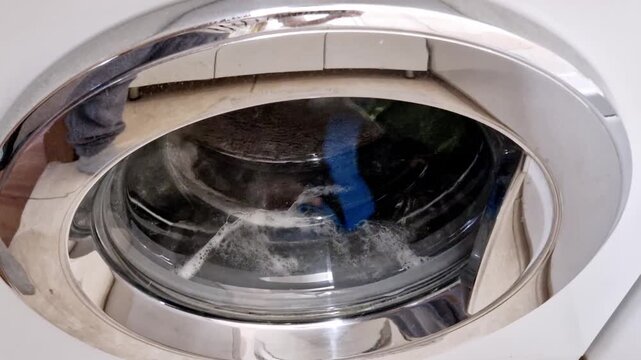 Close-up view from a front-loading washing machine door. The drum is spinning, cleaning colorful clothes with water and white foam bubbles during a regular household laundry cycle in a home.
