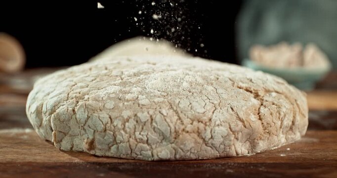 Static shot of a leavened dough loaf isolated on black background with falling flour in super slow motion, 1000 fps. Concept of baking, artisan bread making and dough preparation.