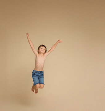 Full length image of a boy at school jumping up with raised hands, dressed only in jeans shorts, and bare feet, isolated beige background.