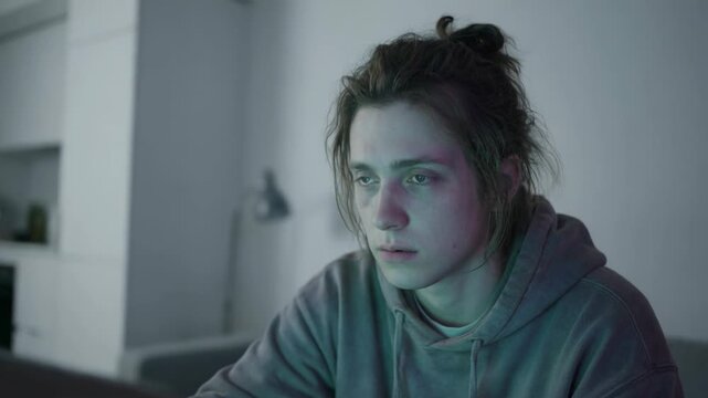 A tired, sleepy young man sits at a table and works on a computer, demonstrating despair and indifference
