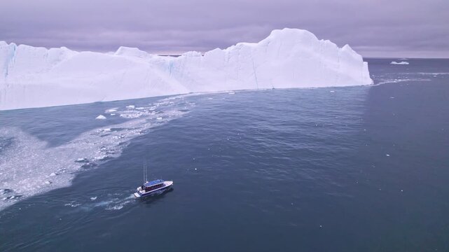 Aerial view of tourist boat sailing icy waters past massive icebergs and floating ice in the Disko Bay, near Ilulissat, showcasing the unique Arctic landscape and a popular travel destination