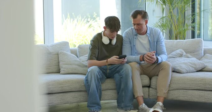 Father and teenage son leaning while son scrolling smartphone on sofa in living room sharing smiles