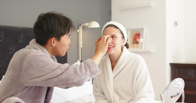 Diverse couple man reaching dabbing cream for skincare placing blue undereye patches on bed in robe