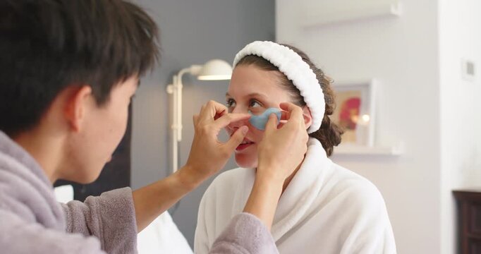 Asian man applying blue under-eye patches on African American woman in plush bathrobes for skincare