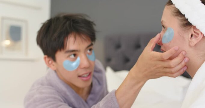 Diverse couple sitting in bedroom after applying under-eye patches, man leaning adjusting her patch