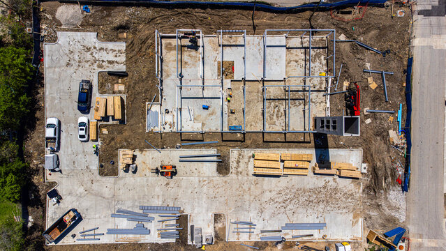 Aerial view of construction site with concrete foundations, lumber stacks, and heavy equipment amidst earthy tones and raw textures, Frisco, Texas, United States.