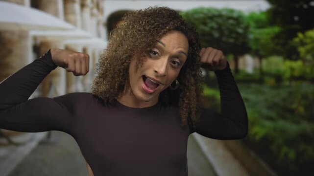 Transgender woman flexes biceps before stone columns of a building with lush green park hedges; confidence.