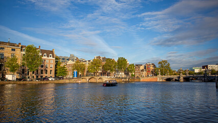 Obraz premium Wide Amsterdam canal with bridge and skyline. Panoramic Amsterdam canal scene with historic buildings, trees, small boat on the water, and a bridge beneath a bright blue sky.