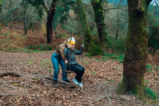 Two happy sisters having fun and laughing while swinging on a wooden swing in the autumn forest.