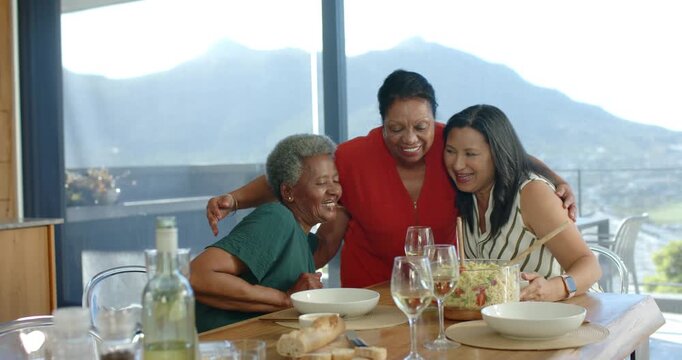 Diverse senior women hugging, center woman reaching, leaning over table sharing meal with salad