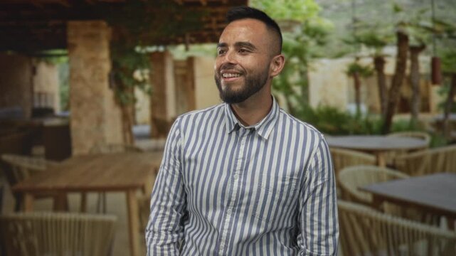 Man with beard smiling, head tilted and looking up, standing by a wooden table in a restaurant building terrace; relaxed joy.