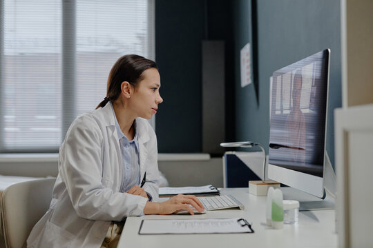Caucasian young adult woman dermatologist consulting male patient online during virtual appointment, sitting at computer desk in medical office setting with documents and clipboard visible