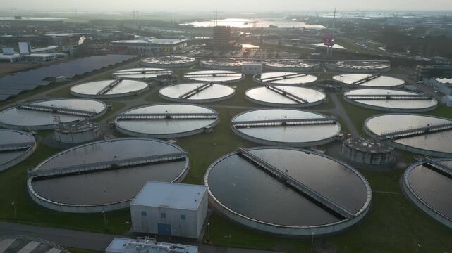 Cinematic drone approach flying over symmetrical circular wastewater clarification tanks at the Harnaschpolder treatment facility in the Netherlands during soft sunrise light.