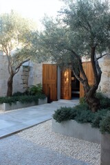 Two majestic olive trees stand gracefully beside a winding path that leads up to open wooden doors, inviting you into a serene courtyard adorned with pebbles and vibrant plants.