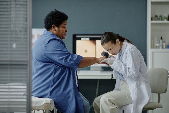Young Asian woman dermatologist examining hand of middle aged Black woman using dermatoscope in medical office, focusing on skin evaluation during dermatology consultation