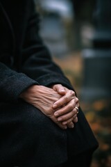 Gently folded hands of older woman in black wool coat resting on lap, funeral concept, grief