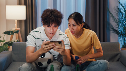 Asian new marriage couple sit on sofa excited cheering watching football sport game on mobile phone in living room at home. Young couple sport fans hold football watching online score on smartphone. © tirachard