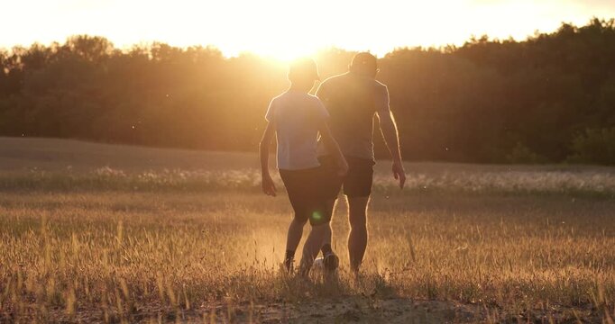 Family spends time playing soccer in countryside field. Father controls ball while son follows movement. Golden sunset, cinematic atmosphere. Happy family, connection, energy and active childhood.