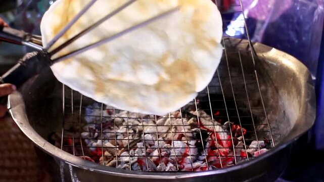 Vendor grilling Khao Krieb Wow giant rice crispy on charcoal grill at Thai night market. Traditional street food snack being flipped with tongs over hot red embers.