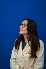 Woman wearing glasses and a coat, contemplating with her head tilted, looking up against a blue background