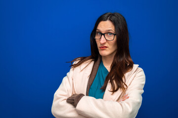 Woman wearing glasses with a skeptical expression, crossing her arms against a solid blue background, signifying doubt and distrust
