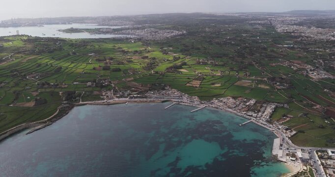 Aerial view of the rugged Munxar Path coastline, where the turquoise sea meets the vibrant green fields and clustered buildings, Marsaskala, Malta.