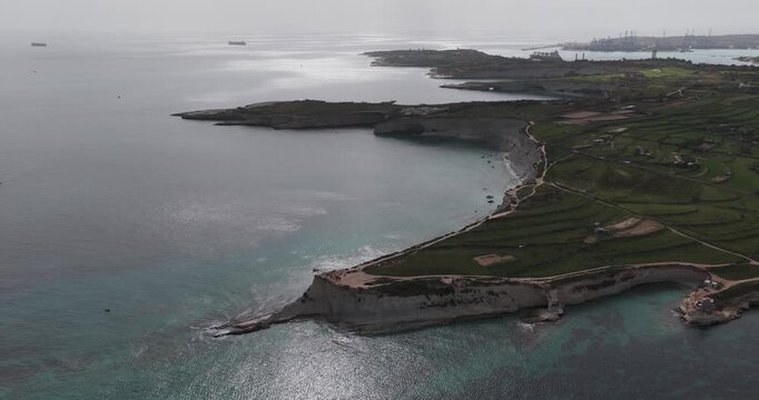 Aerial view of the rugged Malta coastline near Munxar Path, where turquoise waters meet the dramatic cliffs and verdant green fields, Munxar Path, Marsaskala, Malta.