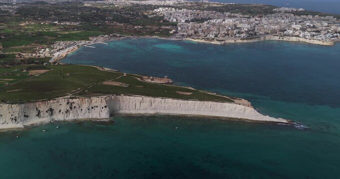 Aerial view of the stark white cliffs meeting the turquoise sea at Munxar Path, with green fields above and the city skyline in the distance, Marsaskala, Malta.