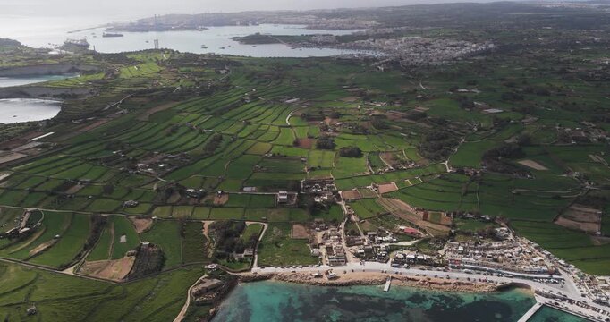 Aerial view of the vibrant green fields contrasting with the rugged coastline and distant urban sprawl, creating a tapestry of natural beauty, Munxar Path, Marsaskala, Malta.